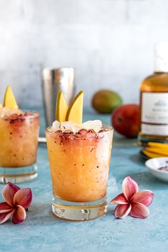 two glasses filled with drinks sitting on top of a blue table next to flowers and fruit