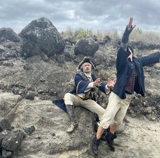 two men dressed as pirates sitting on rocks with their hands in the air and one holding his head