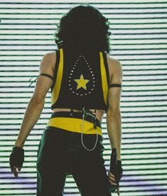 a man with long black hair wearing a yellow and black shirt is standing in front of a screen