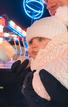 a man holding a baby in front of a carnival ride at night with lights on