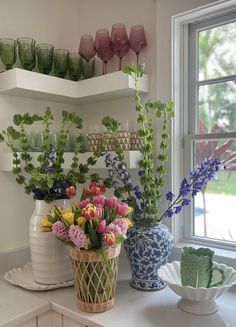 vases filled with flowers sit on a kitchen counter next to plates and cups, near a window