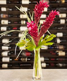 a glass vase filled with red flowers and green leaves next to bottles of wine on the wall