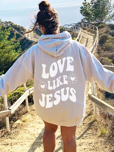 a woman wearing a sweatshirt that says love like jesus on the back, walking down a path