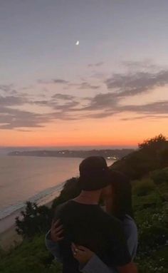 a man holding a woman on top of a hill next to the ocean at sunset