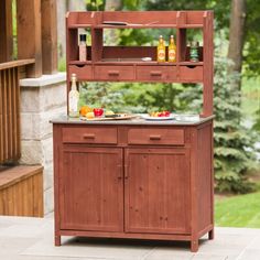 an outdoor kitchen with wooden cabinets and counter tops, outside on a patio in front of some trees