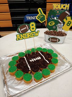 cupcakes with green frosting and brown icing are on a table in front of a football themed cake