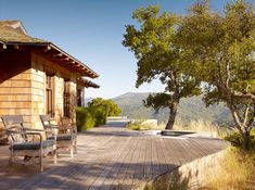 a wooden deck with two chairs on top of it next to trees and mountains in the background