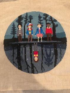 a cross stitch pattern with four people standing on a bridge over a lake in the woods