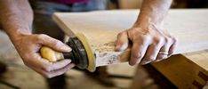 a man sanding wood with a grinder on his right hand and holding a piece of wood in the other hand