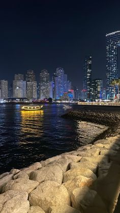 the city skyline is lit up at night with boats in the water and rocks on the shore