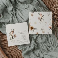 two wedding cards sitting on top of a wicker basket next to dried flowers and greenery