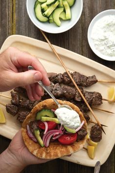 a person holding a skewer with meat and vegetables on it next to bowls of cucumbers