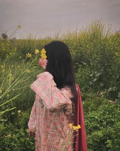 a woman with long black hair standing in the middle of a field holding onto yellow flowers