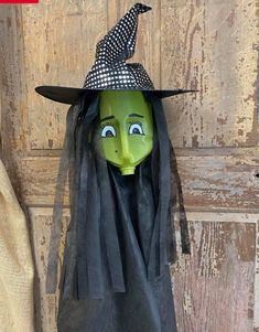 a green witch mask with black hair and a hat on it's head, sitting next to a wooden door