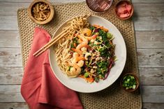 a white plate topped with noodles and shrimp next to chopsticks on top of a mat