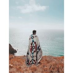 a man standing on top of a grass covered hillside next to the ocean with a blanket over his head