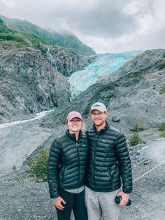 a man and woman standing in front of a glacier with blue water behind them on a cloudy day