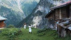 two women are walking in the grass near some mountains and houses, one is wearing a long white dress