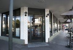 an empty sidewalk with tables and umbrellas in front of a building that has glass doors