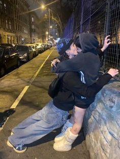 two people kissing each other on the side of a city street at night with cars parked in the background