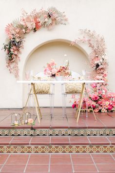 a table set up with pink flowers and greenery in front of a white wall