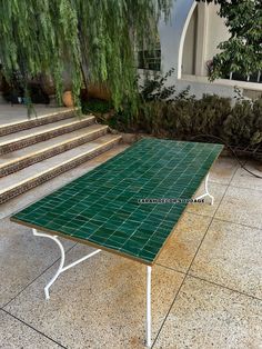 a green table sitting on top of a tiled floor next to some stairs and trees