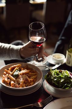 a person holding a glass of wine over a plate of pasta and salad on a table