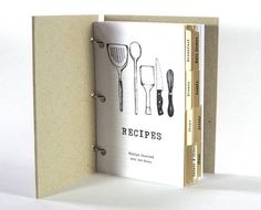 an open recipe book with utensils on the front and back cover, sitting on a white surface