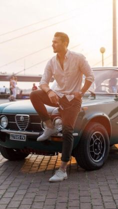 a man sitting on the hood of an old car in front of a bridge at sunset