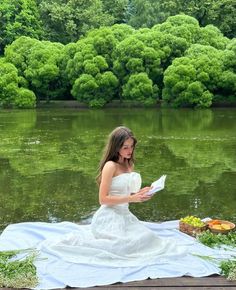 a woman in a white dress sitting on a blanket by the water reading a book