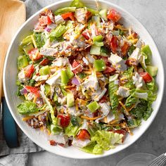 a salad with chicken, lettuce and tomatoes in a white bowl next to a wooden spoon