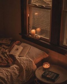 an open book sitting on top of a bed in front of a window with candles