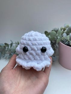 a hand holding a small white crocheted object next to a potted plant