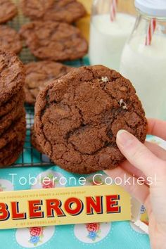 a person holding a chocolate cookie in front of some cookies on a cooling rack and milk