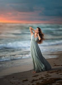 a woman standing on top of a sandy beach next to the ocean at sunset with her hair blowing in the wind