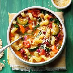 a bowl of pasta and vegetable soup on a green table with two bowls of crackers