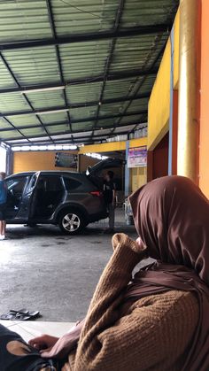 a woman sitting in front of a car under a garage with another person standing next to her