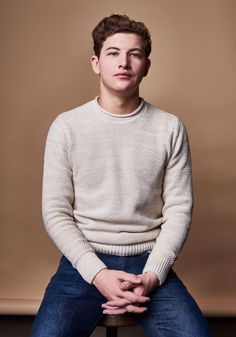 a young man sitting on top of a wooden bench wearing a sweater and jeans with his hands in his pockets