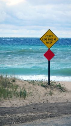 a yellow and red road sign sitting on top of a sandy beach next to the ocean