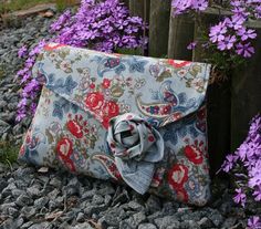 a blue and red flowered purse sitting on top of a pile of rocks next to purple flowers