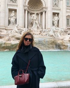 a woman standing in front of a fountain with a handbag on her shoulder and wearing sunglasses