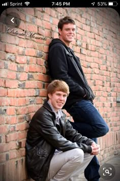 two young men leaning against a brick wall, one is wearing a jacket and the other is smiling