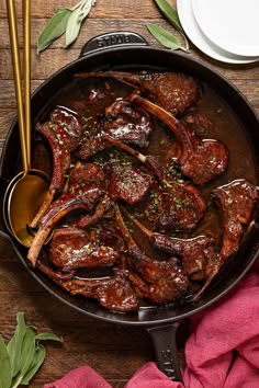 a skillet filled with meat and vegetables on top of a wooden table next to utensils