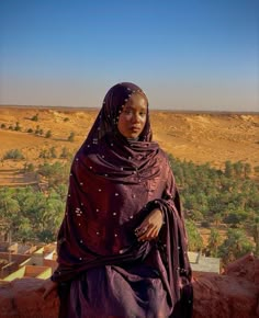 a woman wearing a purple shawl standing in front of a desert landscape with trees