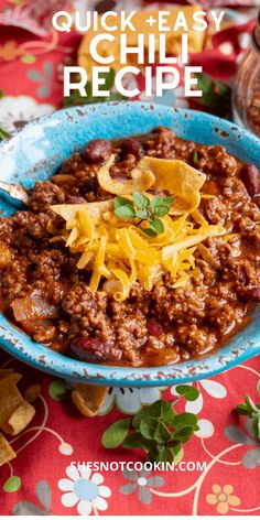 a blue bowl filled with chili and cheese on top of a red table cloth next to a