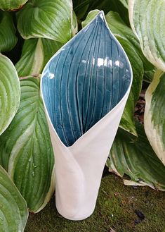 a white and blue vase sitting in front of some green leaves on the ground next to plants