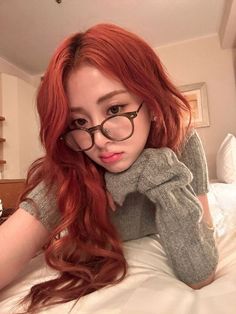 a woman with long red hair and glasses laying on a bed looking at the camera