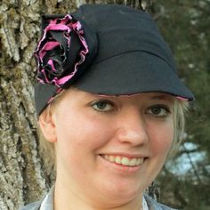 a woman wearing a black hat with a pink flower on the brim, standing next to a tree