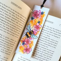 a bookmark with flowers and a bee on it sitting next to an open book