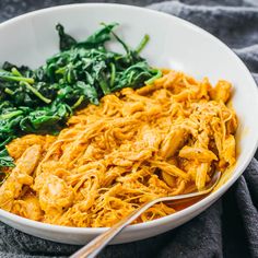 a white bowl filled with pasta and spinach on top of a gray cloth next to a fork